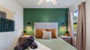 A neatly made bed with pillows and a blanket, a bedside table with a lamp, and a view of greenery through a window with curtains.