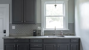 A kitchen with grey cabinets, a white door, a window with a view, a sink, and a countertop with a coffee maker and a soap dispenser.