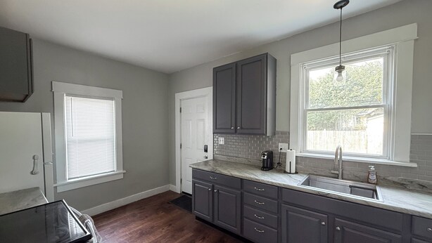 A kitchen with grey cabinets, a marble countertop, and a window with blinds.