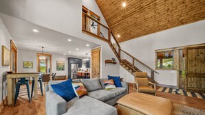 Un vaste espace de vie comprenant un canapé d’angle gris, un pouf marron et un escalier en bois.