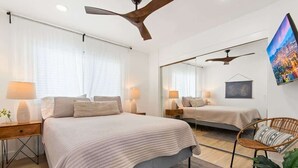 Moderni hotellihuone, jossa on suuri sänky, kaksi yöpöytää, kattotuuletin, suuri peili ja televisio, josta näkyy kaupunkimaisema.