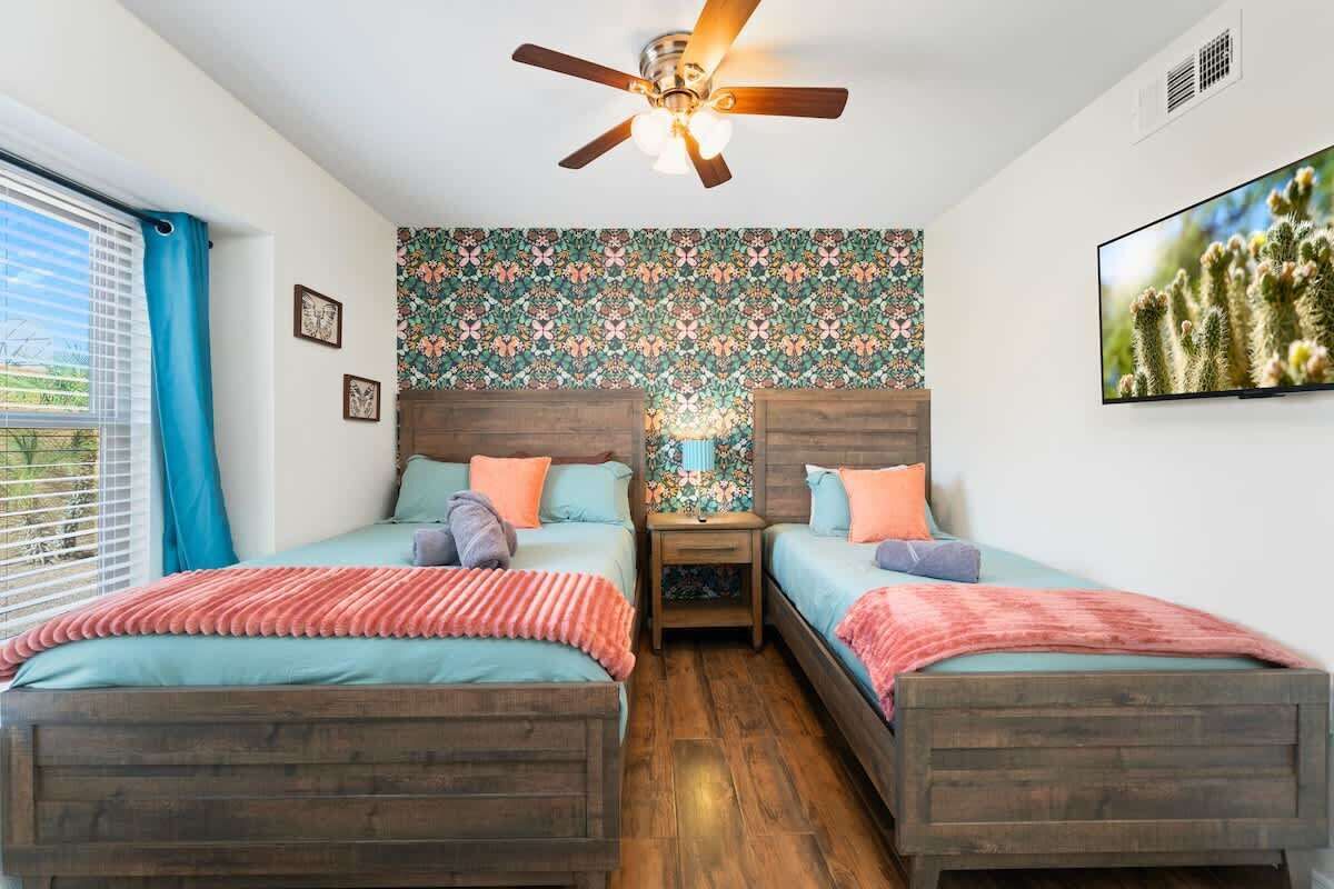 Une chambre à coucher avec deux lits en bois, un ventilateur de plafond, un téléviseur fixé au mur et une fenêtre avec des rideaux.