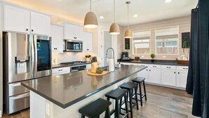 Une cuisine moderne dotée d’appareils électroménagers en acier inoxydable, d’un grand îlot central avec des tabourets, d’un éclairage suspendu et de meubles de rangement blancs.