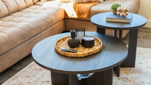 Un salon avec un canapé en cuir marron, deux petites tables rondes, un plateau en osier contenant un vase et une télécommande, et une petite plante en pot posée sur une table d’appoint.