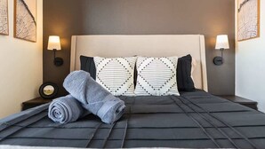Ein ordentlich bezogenes Bett mit einem schwarz-weißen Kissenmuster, einem aufgerollten grauen Handtuch und einem Kopfteil.