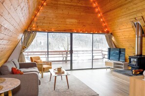 A cozy loft-style room with a wooden ceiling, a large window, a sofa, a TV, and a wood-burning stove.