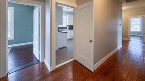 A spacious room with hardwood flooring, white walls, and a door leading to a kitchen area.