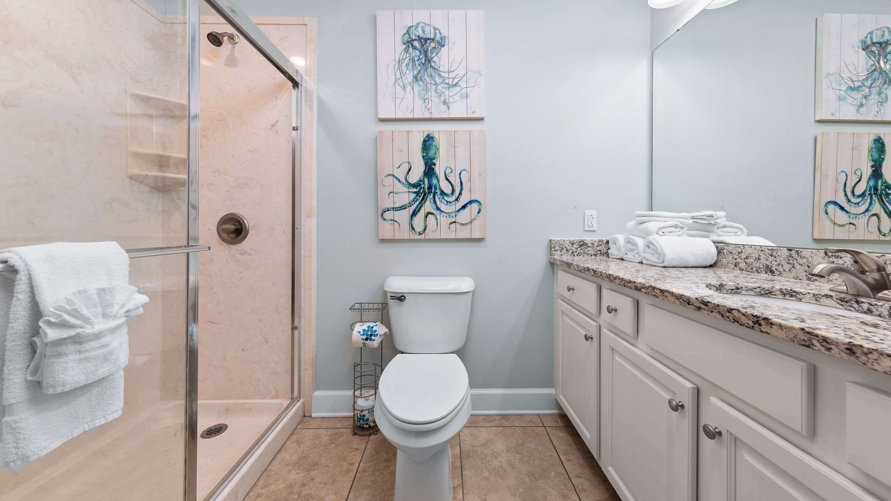 A bathroom with a glass shower enclosure, a toilet, and a vanity with a sink. There are three wall-mounted artworks depicting octopuses.