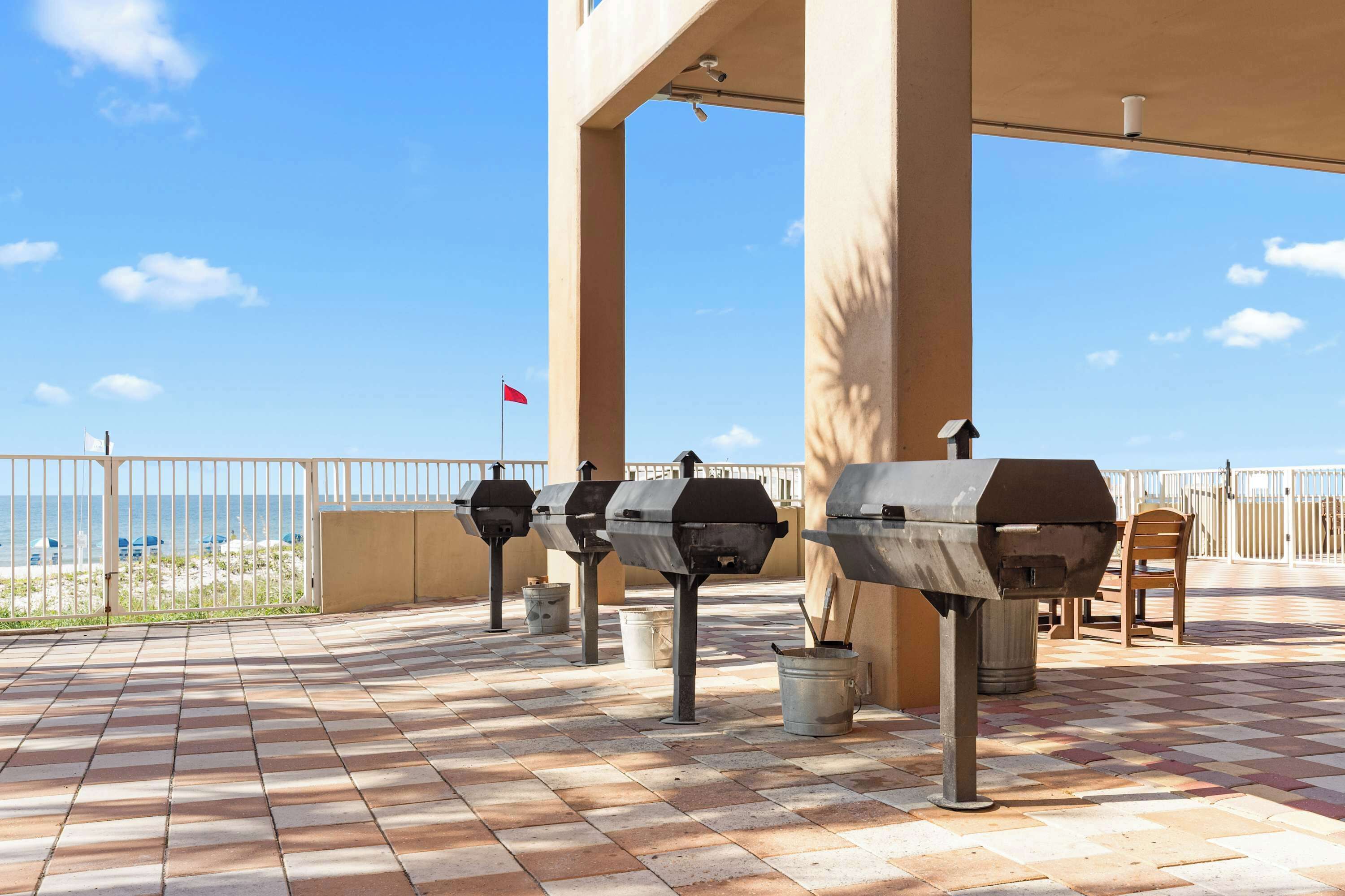 Un patio exterior con parrillas de barbacoa, vista a la playa y una bandera al fondo.