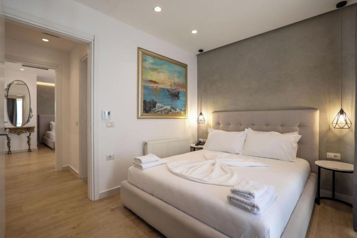 A modern bedroom with a large bed, a painting on the wall, and a view of the sea.