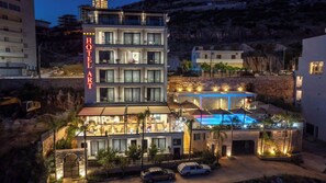 Un hotel de varios pisos con zona de piscina, rodeado de otros edificios y montañas al fondo.
