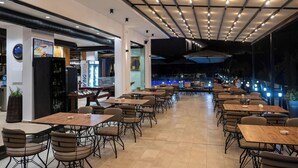 Un'ampia sala da pranzo interna con diversi tavoli e sedie, un angolo bar con frigorifero e vista su una piscina esterna.