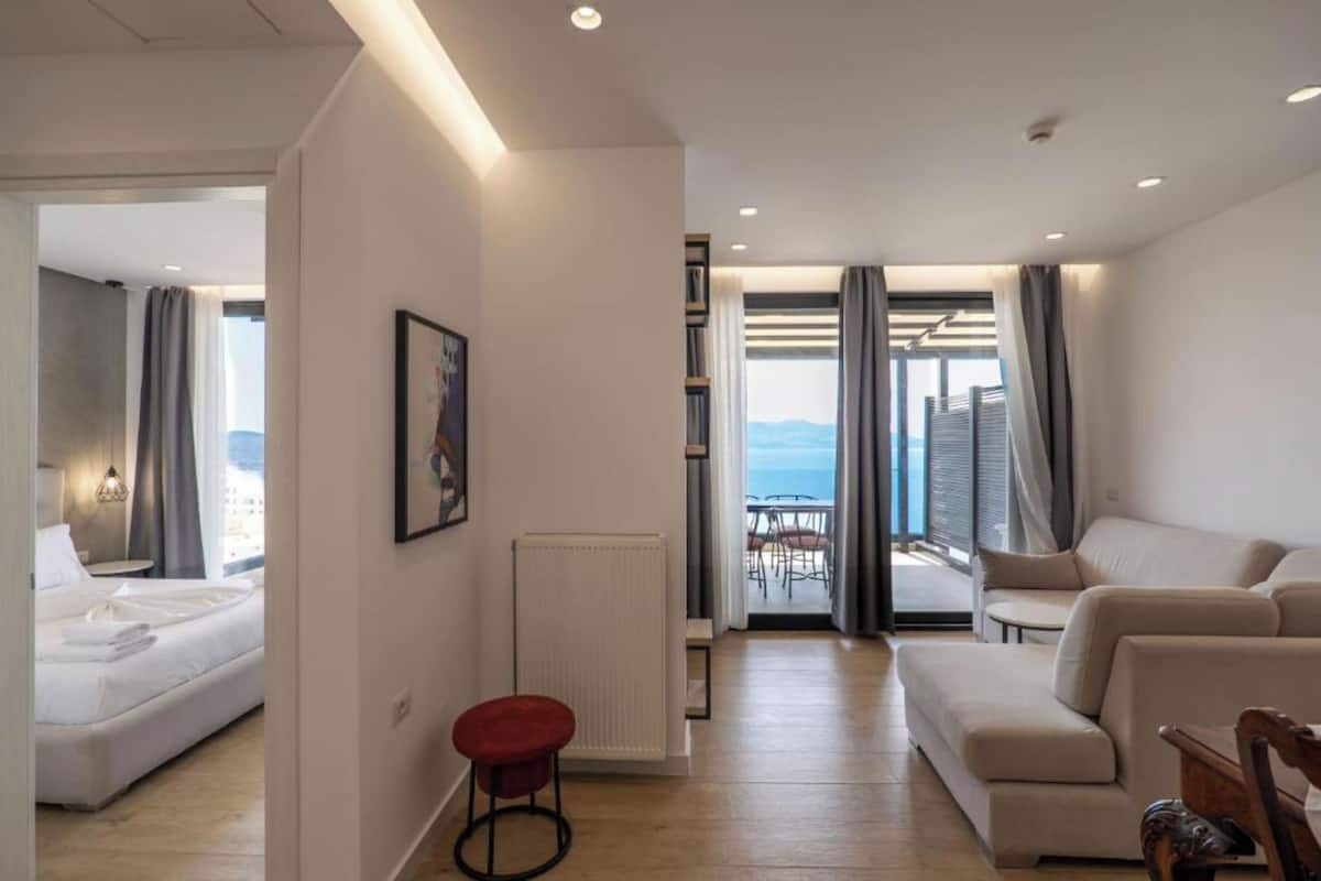 A modern hotel room with a bed, a red stool, a sofa, and a view of the sea through large windows.