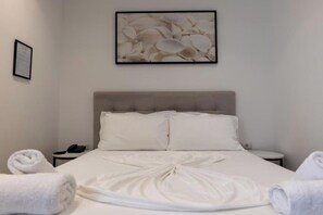 Una cama bien hecha con sábanas blancas y una toalla, una obra de arte enmarcada encima, y una mesita de noche con un teléfono.