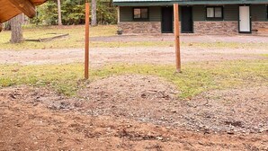 Een rustieke houten constructie met een metalen dak, een stenen gebouw met een groen dak op de achtergrond, en een grasveld ervoor.