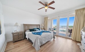 A bedroom with a large bed, a nightstand, a lamp, a ceiling fan, and a view of the outside through large windows.