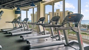 Un gimnasio con cintas de correr y equipamiento de ejercicio, amplias ventanas con vistas a edificios y al océano.