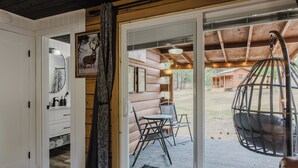 L’intérieur d’une cabane en rondins, avec une porte-fenêtre coulissante donnant sur un patio extérieur équipé d’un fauteuil suspendu et d’une table.