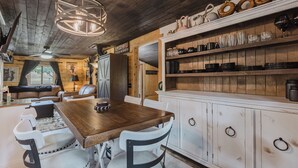 Un coin repas rustique avec une table et des chaises en bois, un lustre et des étagères où sont disposés de la vaisselle et des verres.