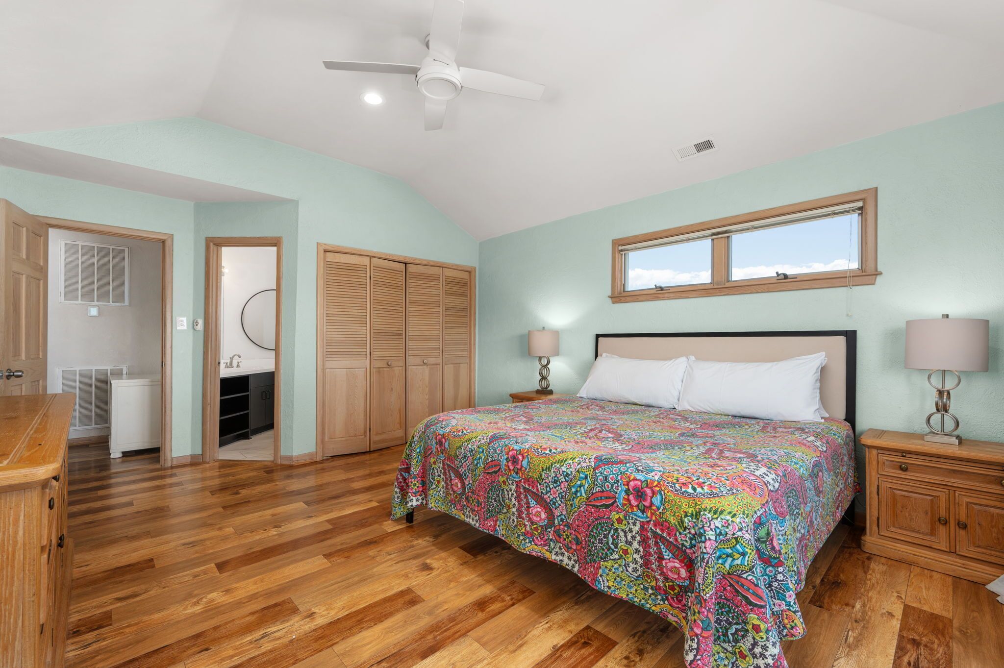 A bedroom with a large bed, wooden floor, a ceiling fan, and a window with a view.