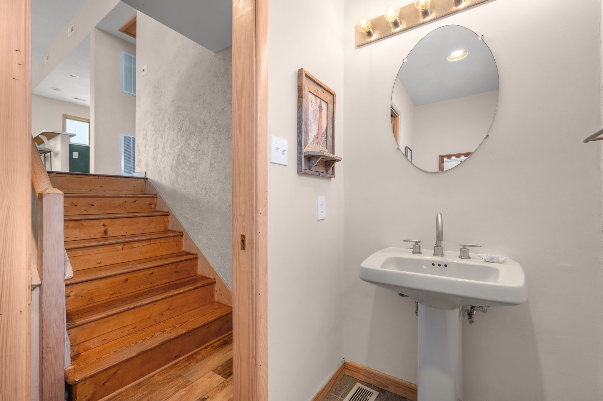 Un escalier en bois mène à une salle de bain  équipée d’un lavabo blanc et d’un miroir rond.