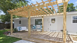 Een houten pergola met een terras, een witte deur en een kleine tuin met planten en een bank.