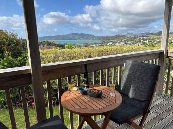 A wooden deck with a round table and two chairs, overlooking a scenic landscape with hills and a body of water. - Casa Playa de los osos | Coopers Beach Sea Views (Cable Bay)
