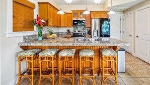 A kitchen with wooden cabinets, a granite countertop, a stainless steel refrigerator, and a bar area with wicker stools.