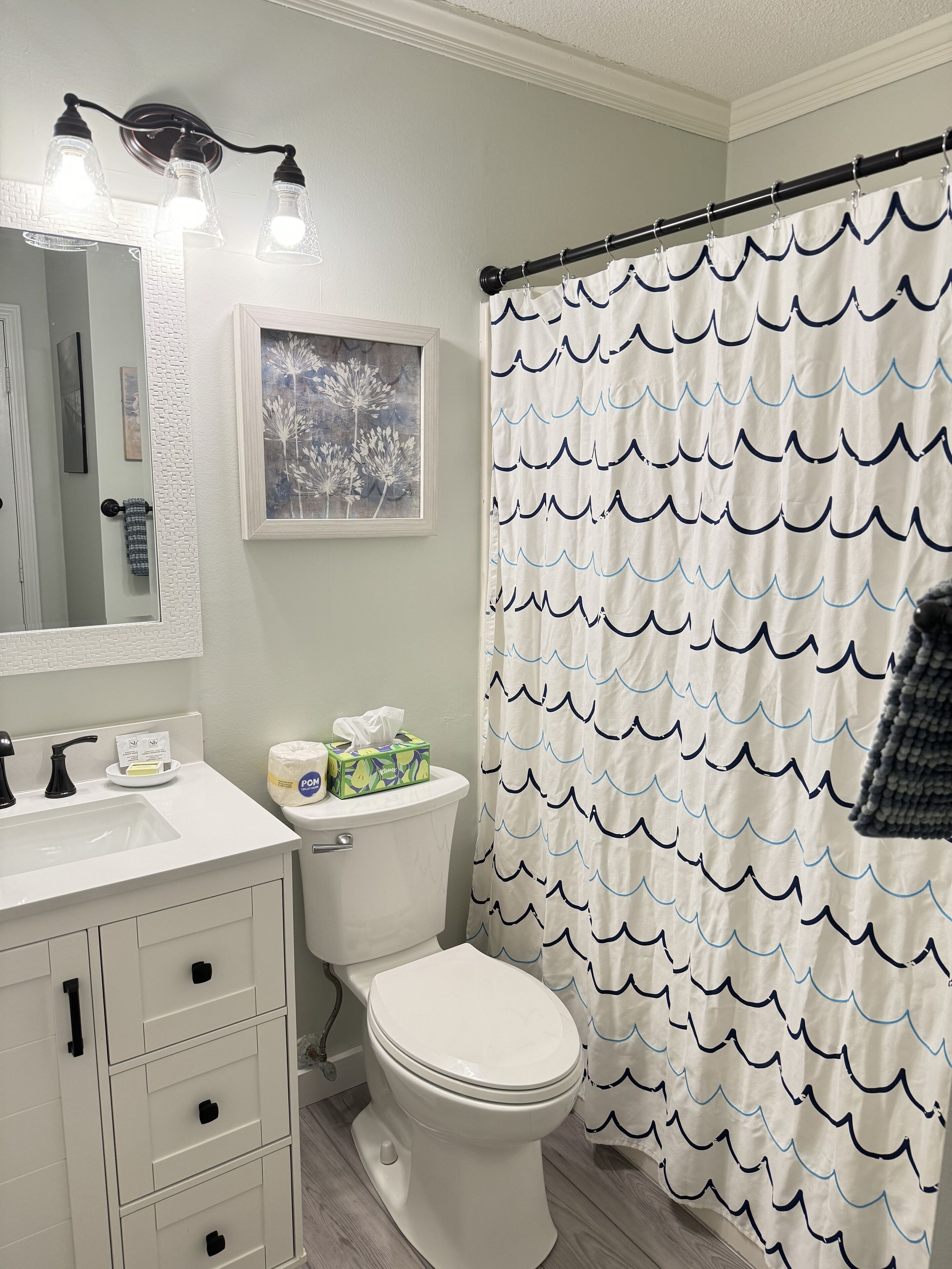 Une salle de bain avec des toilettes, un lavabo et un miroir. Il y a un rideau de douche orné d’un motif de vagues.