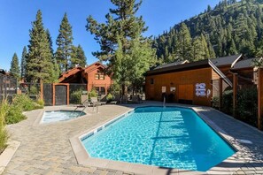 Uma área de piscina com duas banheiras de hidromassagem e cabanas de madeira, cercada por árvores e um cenário montanhoso.