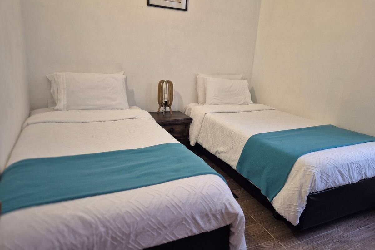 Une chambre avec deux lits, chacun agrémenté de linge de lit blanc et d’un élément décoratif de couleur turquoise. Un tableau encadré est accroché au mur, au-dessus des lits.