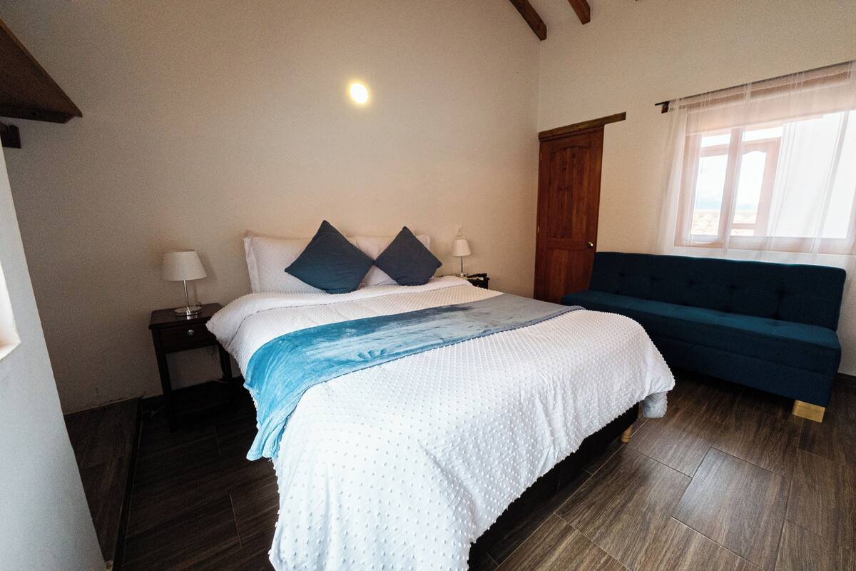Une chambre à coucher avec un lit, une table de chevet, un canapé bleu et une fenêtre avec des rideaux.
