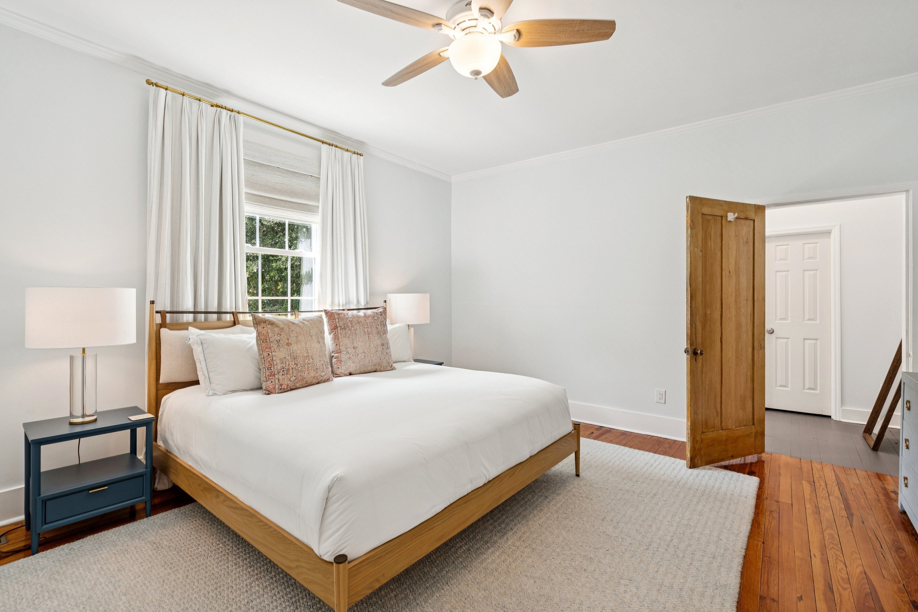 A bedroom with a wooden bed, white bedding, a ceiling fan, a window with white curtains, and a door.