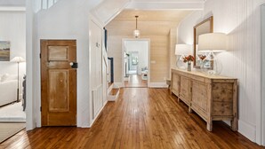 Un hall d’entrée spacieux avec un plancher en bois, une porte en bois, un escalier blanc et une console en bois agrémentée de lampes et d’objets décoratifs.