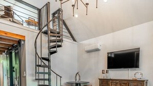 A spacious living room with a spiral staircase, a round coffee table, and a large flat-screen TV. - Private Lakeside Couples Cabin, Hot Tub & Fire Pit (Bullard)