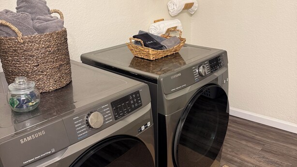 Un área de lavandería con dos lavadoras Samsung, una cesta de mimbre con toallas y un frasco pequeño de vidrio.