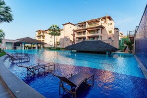 A swimming pool with lounge chairs and a thatched-roof structure, surrounded by multi-story buildings with balconies. - Flat Beverly Hills - Porto das Dunas (Aquiraz)