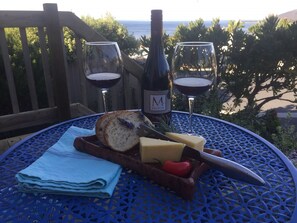A table with a bottle of wine, two glasses, a knife, cheese, and bread on a blue table outdoors with a view of the ocean. - Bicheno Bell a cosy stay for couples and fantastic proximity! (Bicheno)