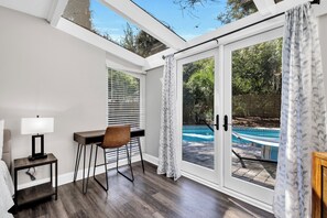A sunlit room with a desk, chair, and a view of a swimming pool through glass doors.