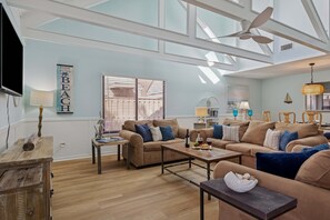 A spacious living room with a high ceiling, large windows, and a ceiling fan. There are two sofas and a coffee table, with a TV and a lamp on the wall.
