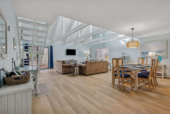 A spacious living area with a high ceiling, a dining table with chairs, and a television mounted on the wall.