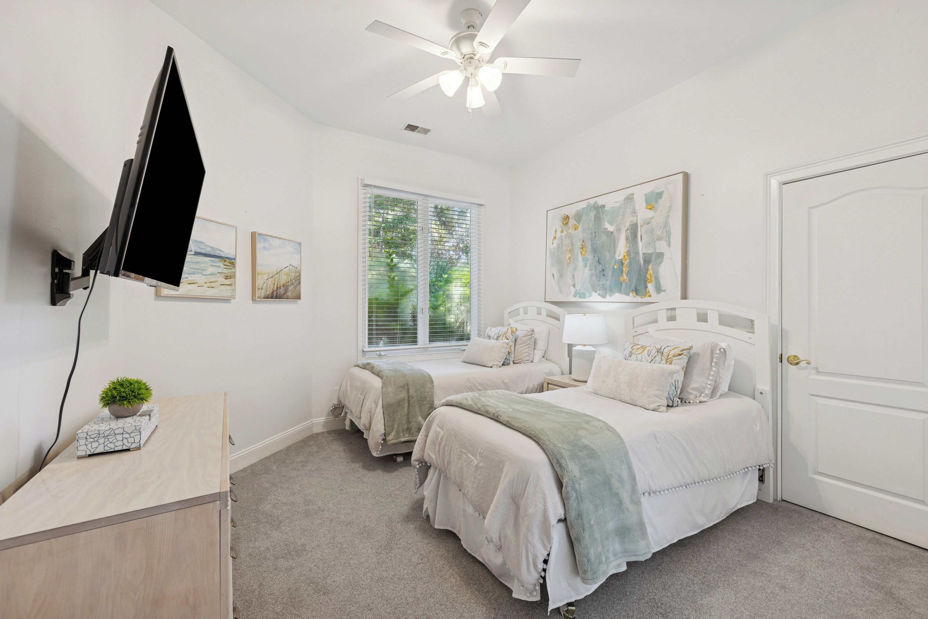 A bedroom with two beds, a ceiling fan, a TV mounted on the wall, and artwork on the walls.