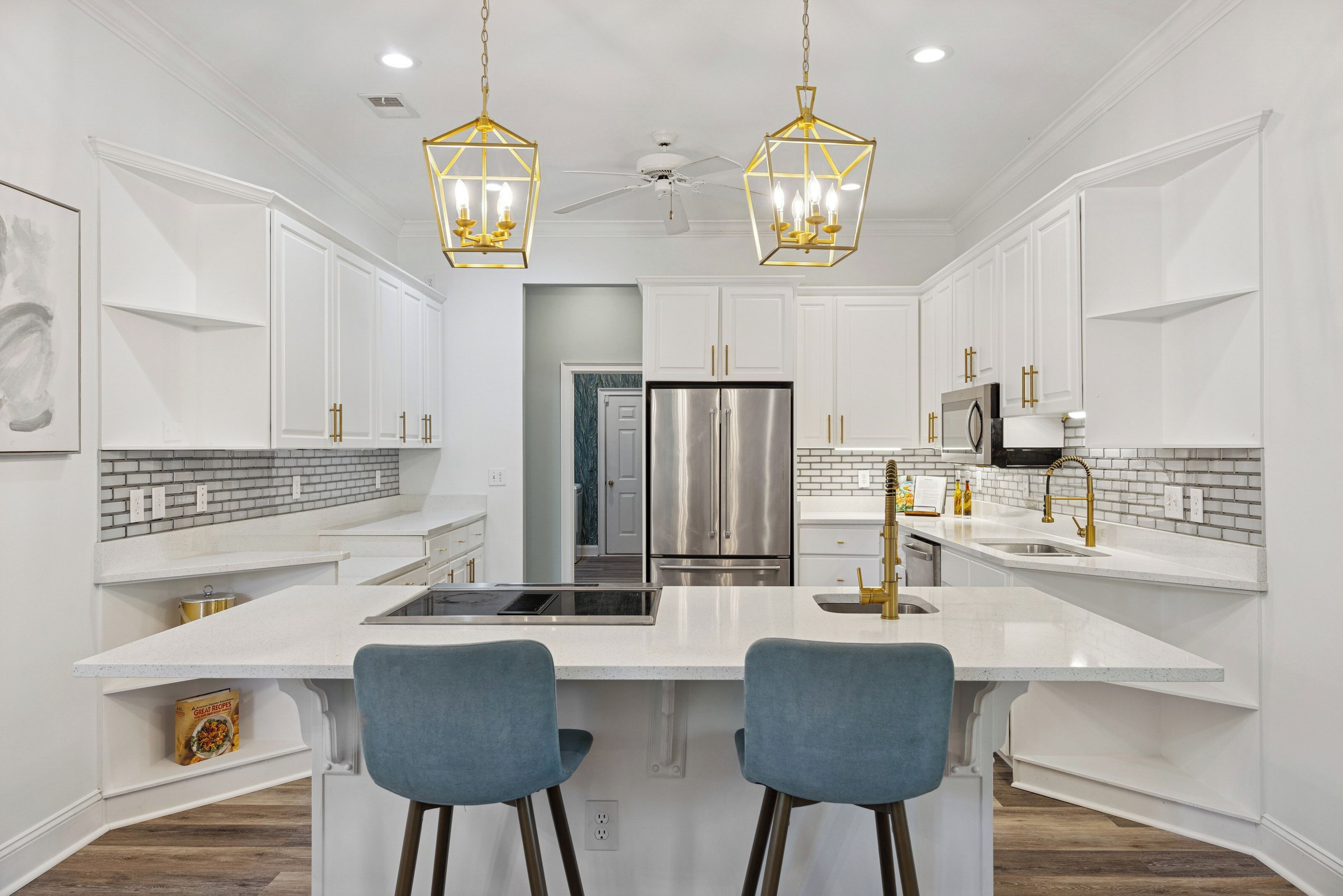 Une cuisine moderne avec des meubles blancs, un îlot central doté de tabourets de bar, des appareils en acier inoxydable et des luminaires suspendus aux tons dorés.