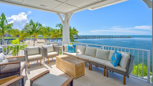 A covered outdoor seating area with wicker furniture and cushions, overlooking a body of water and a pier. - Emerald Shores – Luxury Oceanfront Rental, Heated Pool & Private Dock. Pets Welcome! (Marathon)