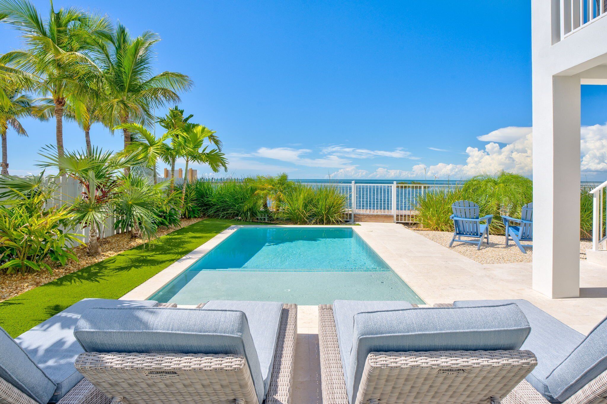 A rectangular swimming pool with clear blue water, surrounded by lush green grass and palm trees. There are two wicker lounge chairs with blue cushions facing the pool.