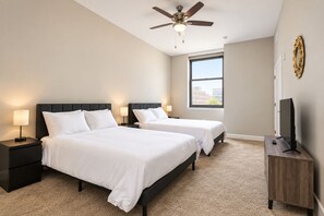 A hotel room with two beds, a ceiling fan, a TV on a wooden stand, and a window with a view of buildings.