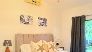 Een slaapkamer met een bed, een plafondventilator, een airconditioningapparaat, een nachtkastje, een lamp en een raam met gordijnen.