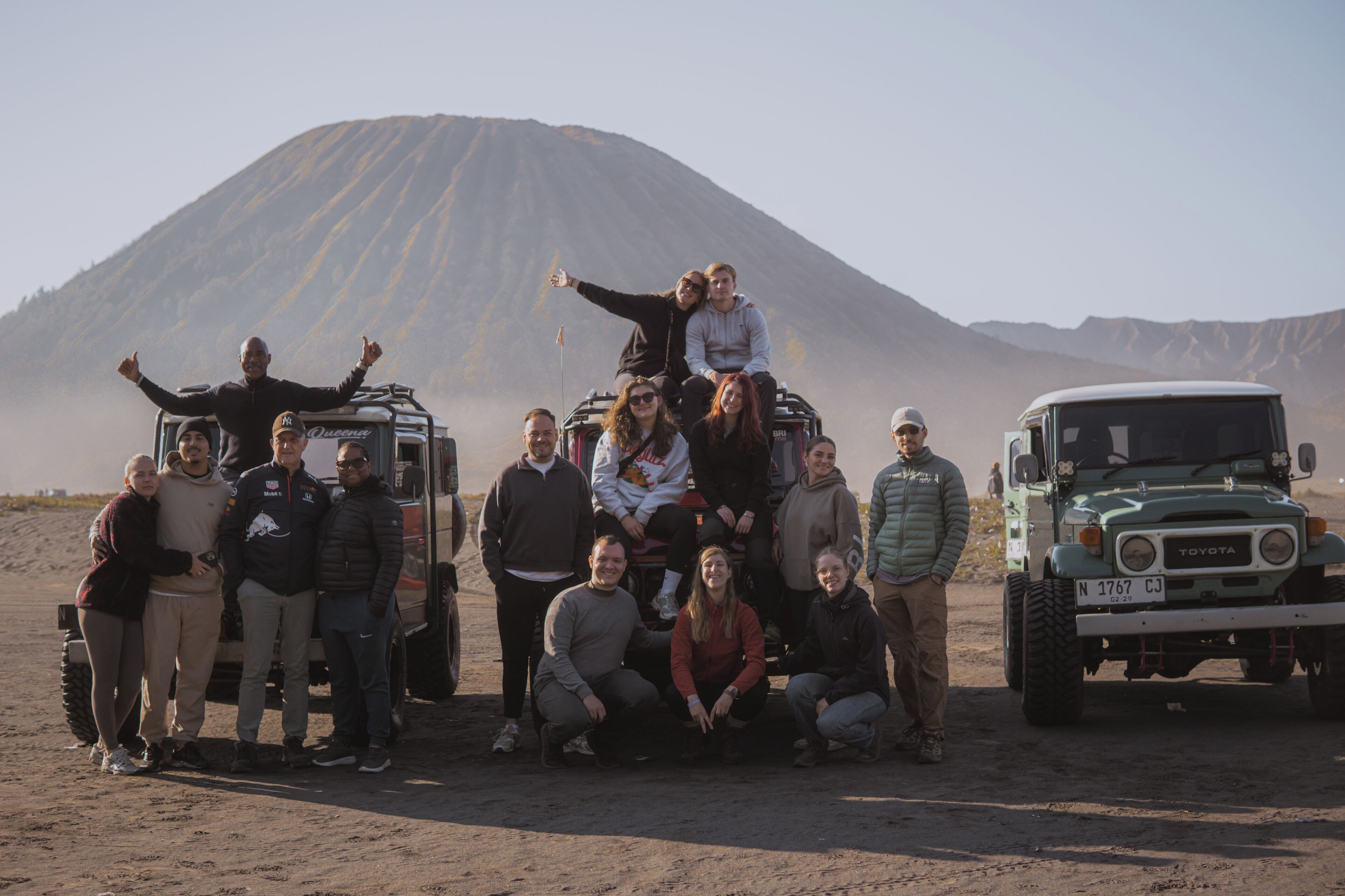 Un grupo de personas posando frente a un vehículo con montañas al fondo.