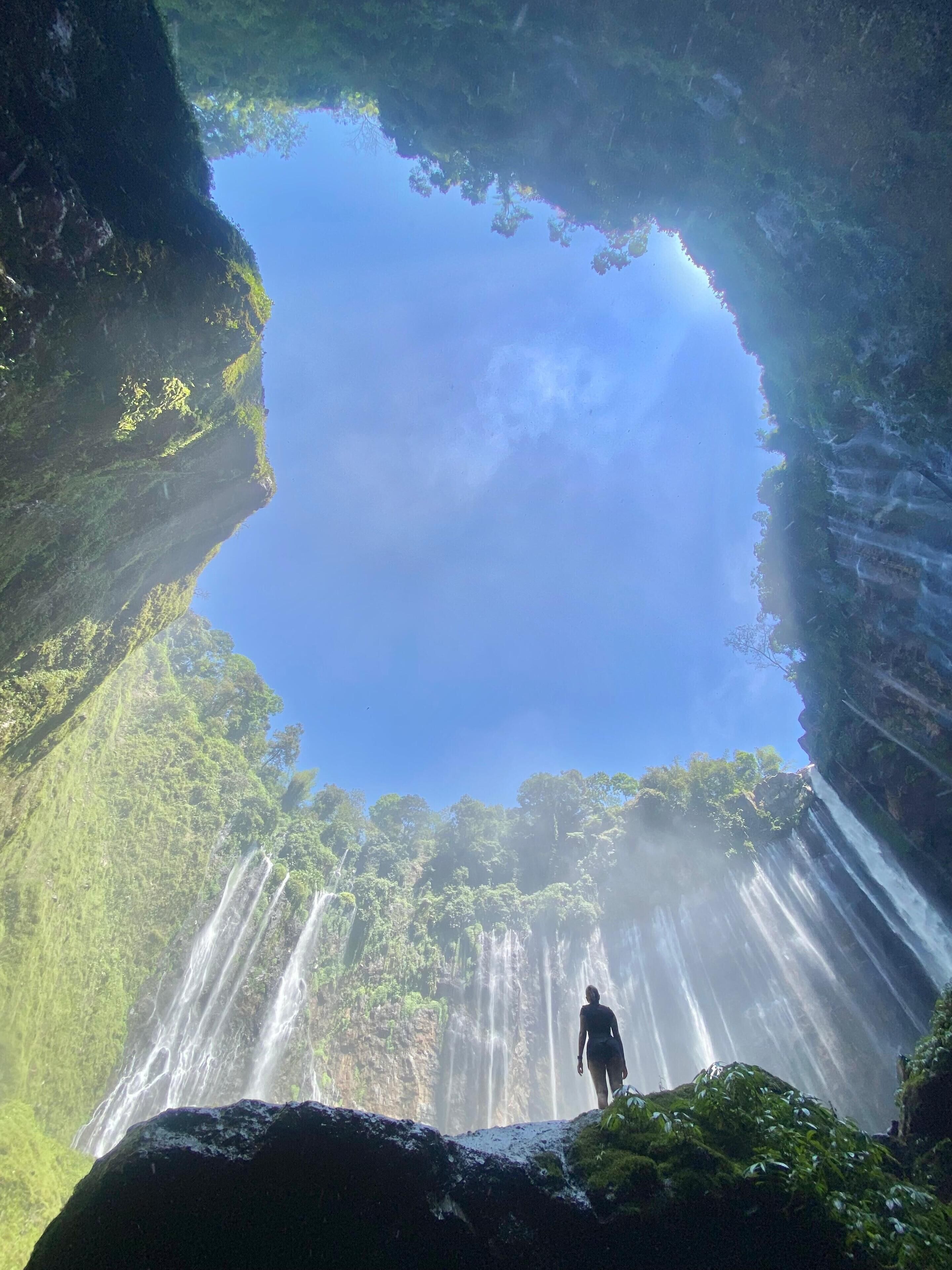 A person standing at the edge of a waterfall with a clear view of the sky above.