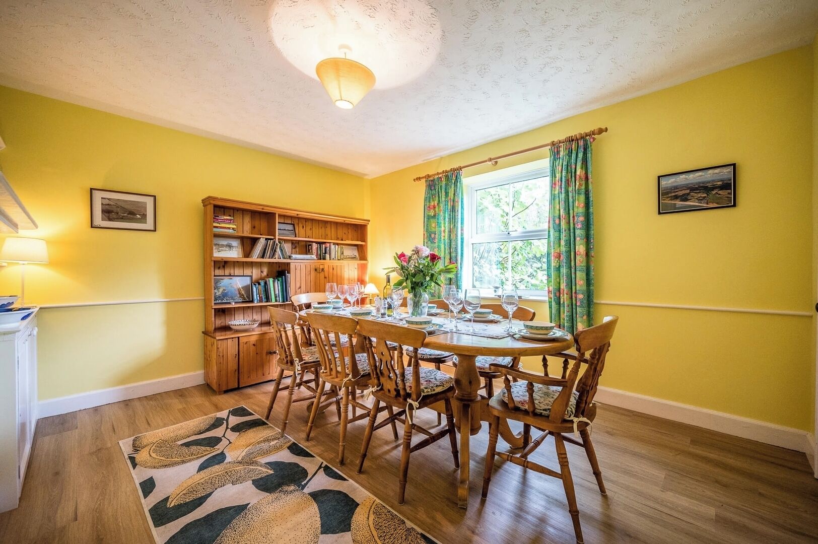 A dining area with a wooden table set for four, surrounded by chairs. A bookshelf filled with books and decorative items is to the left.